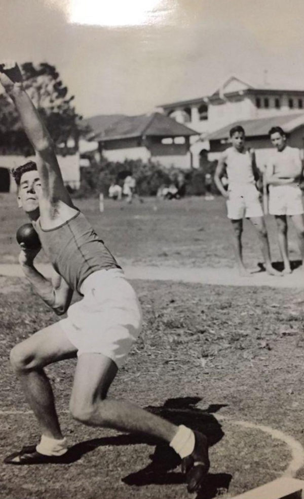 A young Laurence Quinlivan crouched to put the shot, with two athletes watching